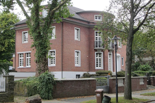 Backsteingebäude mit grauem Dach, weissen Fenstern und Bäumen vor der Fassade.