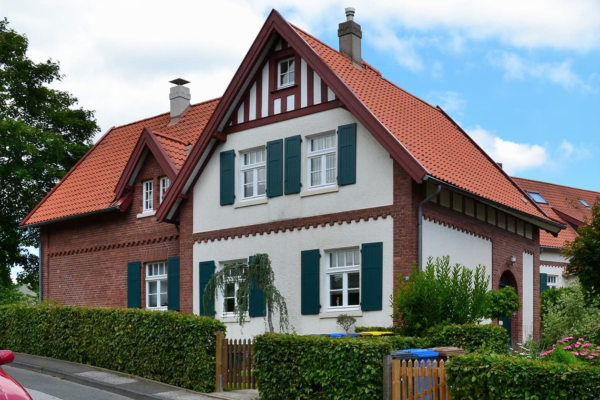 Backsteinhaus mit rotem Ziegeldach, gruenen Fensterlaeden und Hecke vor der Fassade.