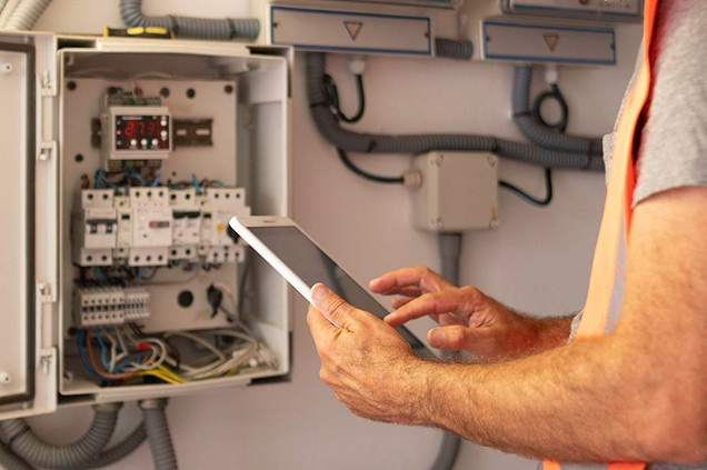 Fachperson prueft einen offenen Schaltschrank mit einem Tablet in der Hand.