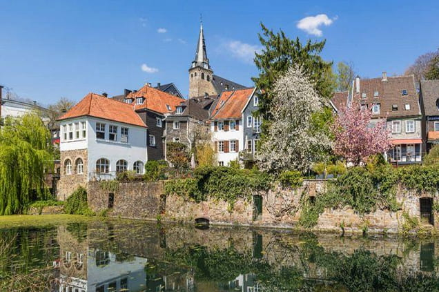 Eine Reihe historischer Häuser mit roten und grauen Dächern steht an einem Teich, im Hintergrund sind unter blauem Himmel blühende Bäume und ein Kirchturm zu sehen.