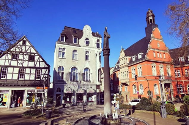 Der Stadtplatz von Essen-Werden mit einer Statue in der Mitte, umgeben von historischen Gebäuden mit einem Fachwerkhaus und einem Gebäude aus rotem Backstein unter strahlend blauem Himmel.