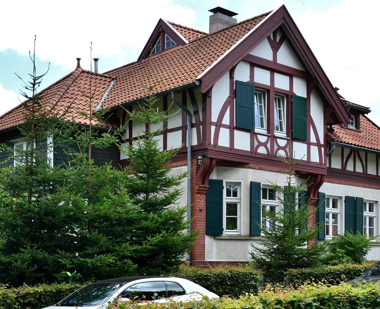 Fachwerkhaus mit rotem Ziegeldach, gruenen Fensterlaeden und Baeumen im Vordergrund.