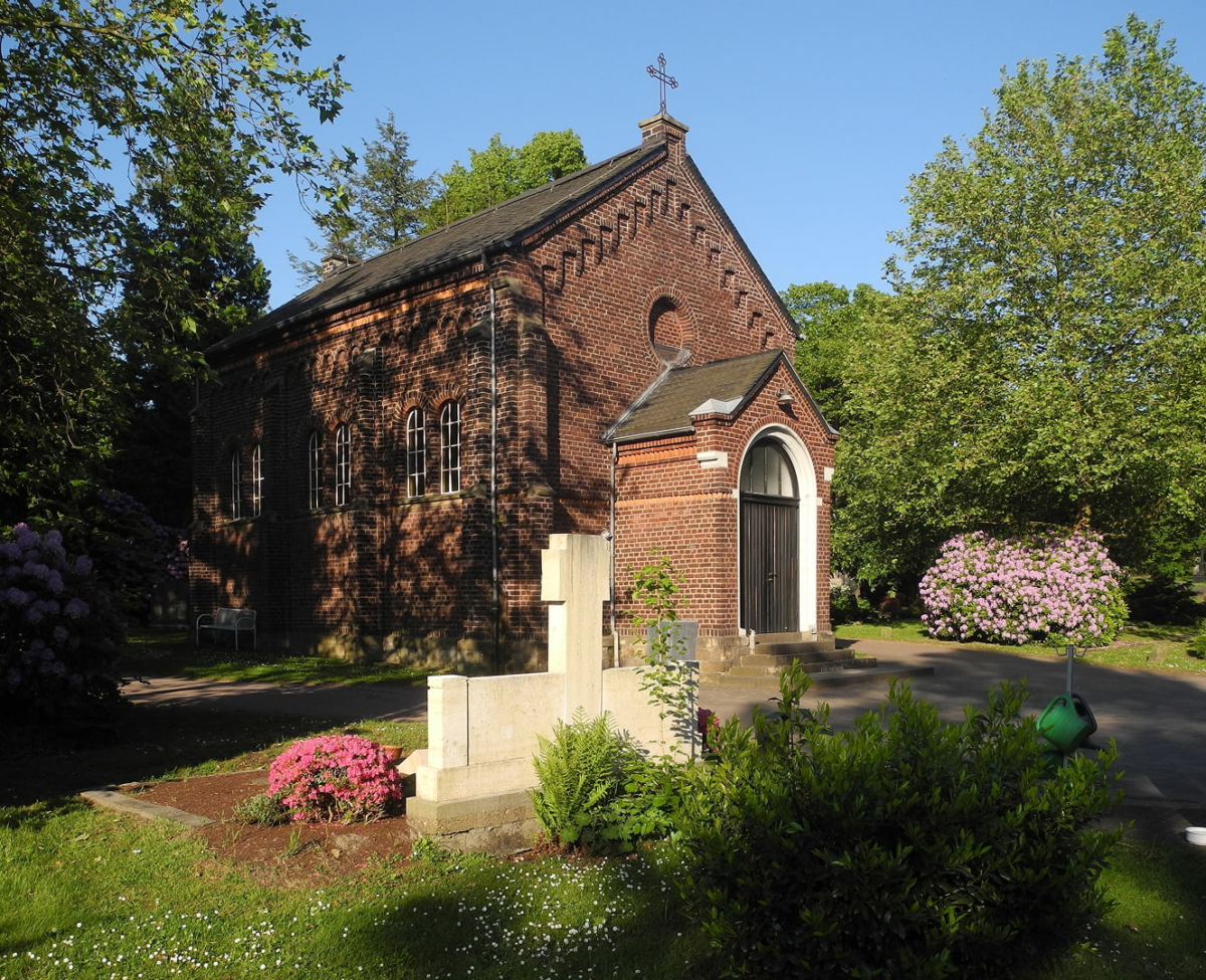 Kleine Backsteinkapelle auf einem Friedhof in Essen-Werden mit blühenden Rhododendren und einem hellen Steinkreuz.