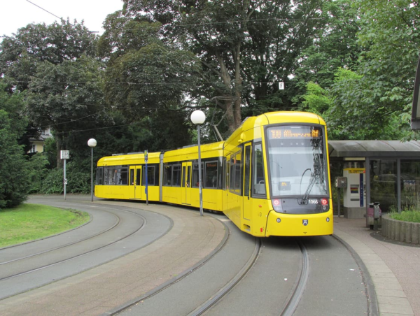 Gelbe Strassenbahn an einer Haltestelle in gruener Umgebung.