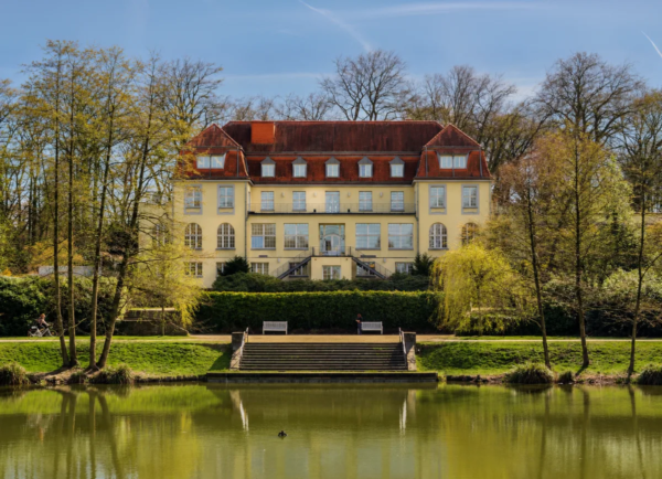 Historisches Herrenhaus mit rotem Dach hinter einem Teich und alten Bäumen in Mülheim an der Ruhr