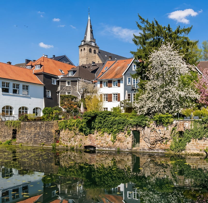 Historische Altstadt mit Kirche, Wohnhäusern und blühenden Bäumen am Wasser.