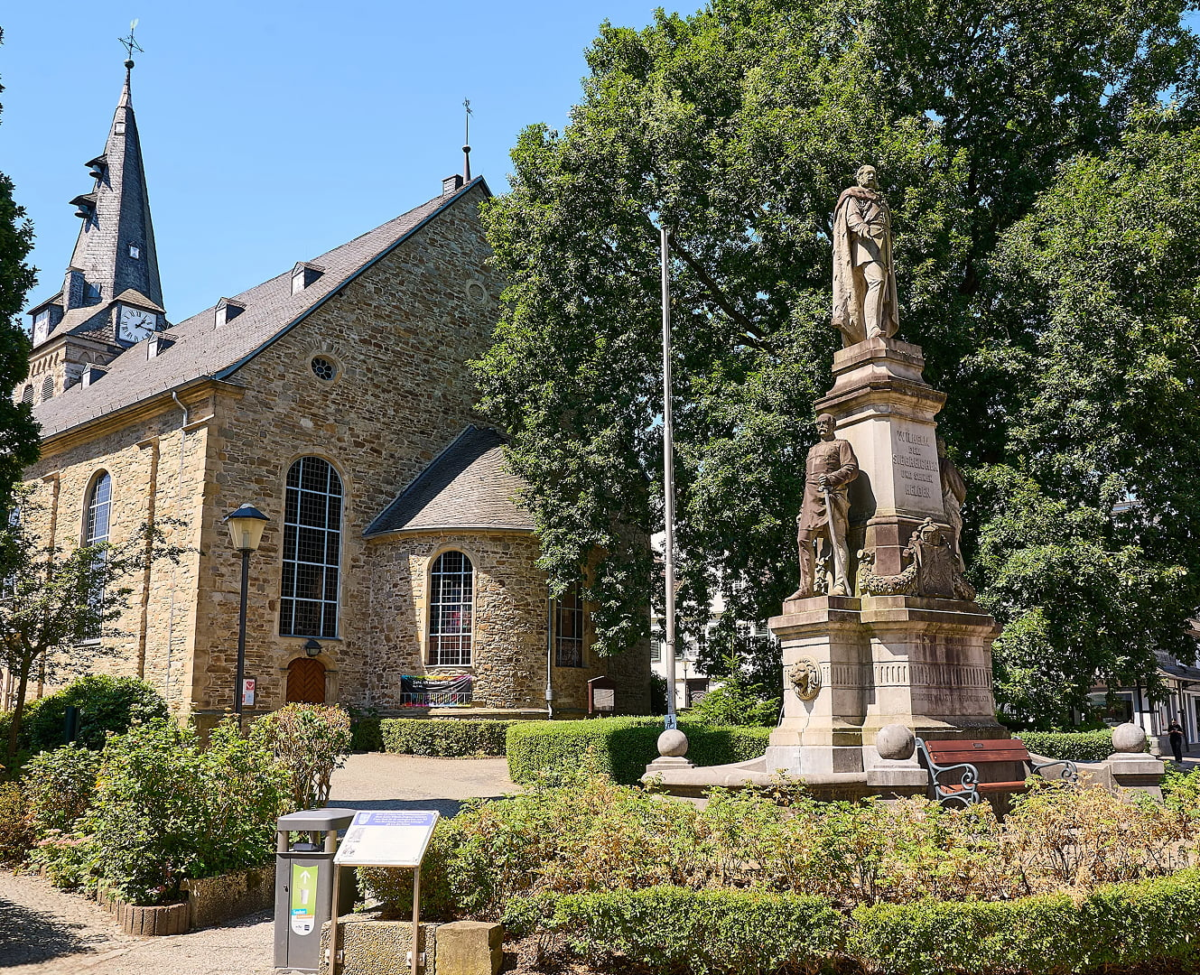 Historische Bruchsteinkirche mit Schieferturm und Kaiser-Wilhelm-Denkmal auf einem begrünten Stadtplatz.