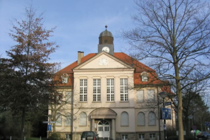 Historisches Gebäude mit Uhrturm und Vorplatz zwischen Bäumen.