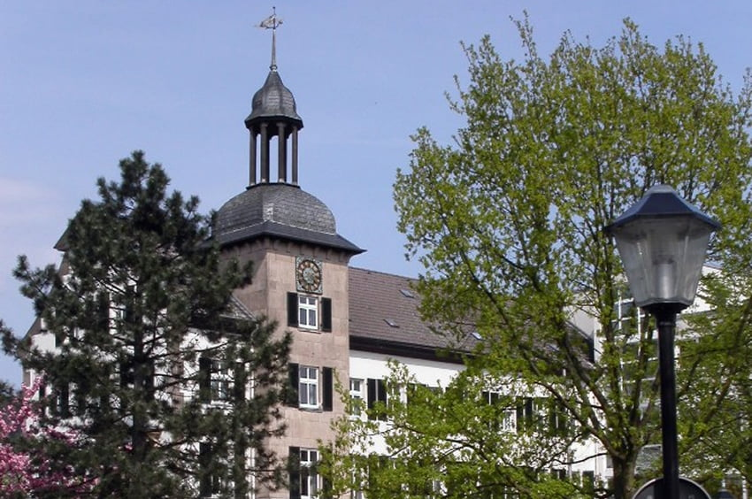 Historisches Gebäude mit Uhrturm, umgeben von Bäumen.