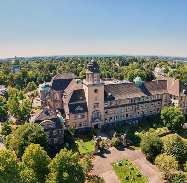 Luftaufnahme eines historischen Schulgebäudes mit Turm, eingebettet in einen grünen Stadtteil.
