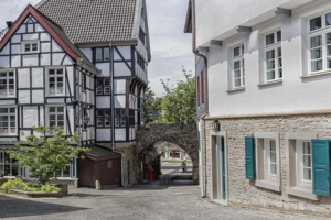 Eine Kopfsteinpflasterstraße schlängelt sich an Fachwerk- und Steinhäusern vorbei und führt zu einem gewölbten Steintor, hinter dem Bäume zu sehen sind.