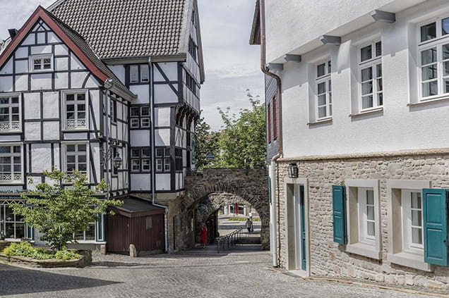 Eine Kopfsteinpflasterstraße schlängelt sich an Fachwerk- und Steinhäusern vorbei und führt zu einem gewölbten Steintor, hinter dem Bäume zu sehen sind.