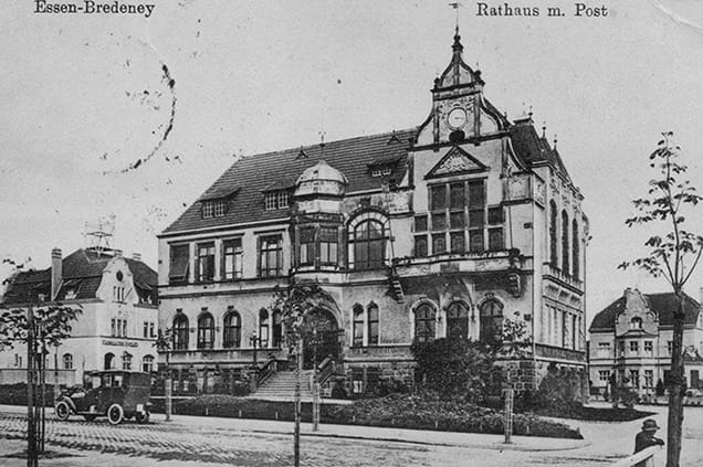 Schwarz-Weiß-Foto des Rathaus- und Postgebäudes in Essen-Bredeney mit einem Uhrenturm und Architektur aus dem frühen 20. Jahrhundert, daneben steht ein Oldtimer.