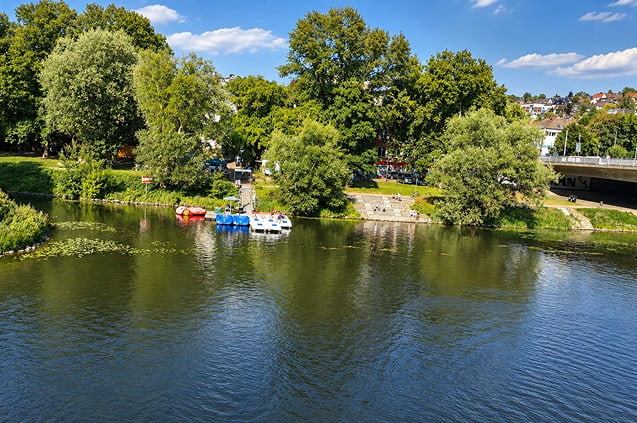 Ruhrufer in Essen-Werden mit bunten Tretbooten, Bäumen und einer Brücke im Hintergrund.