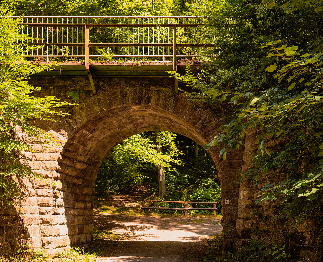 Steinerne Bogenbrücke über einem Waldweg in grüner Umgebung.