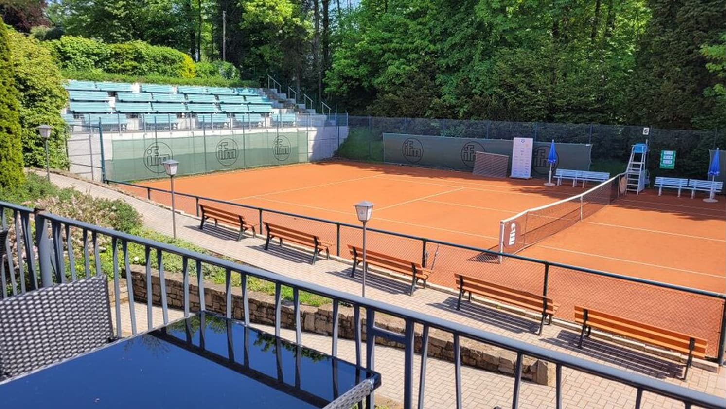 Tennisplatz mit rotem Sandbelag, Netz, Sitzbaenken und Zuschauerbereich am Waldrand.