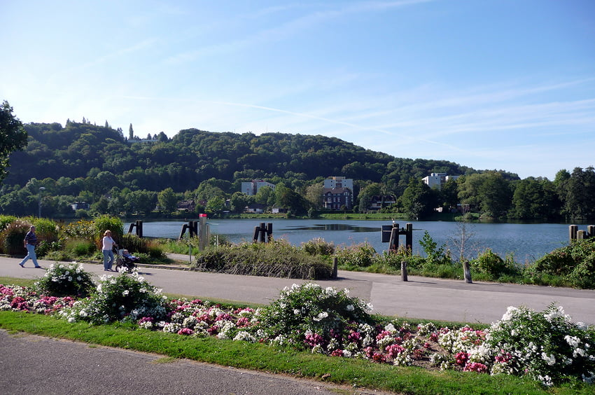 Uferpromenade mit Blumenbeeten und Blick auf ein bewaldetes Gewässer.