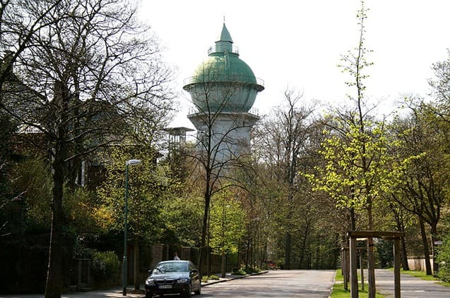 Wasserturm hinter Bäumen an einer ruhigen Strasse mit geparktem Auto.