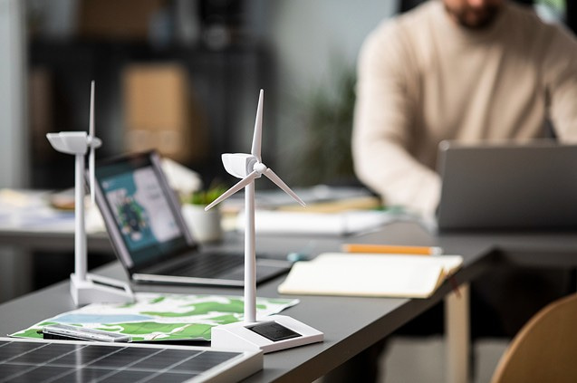 Windradmodell auf einem Schreibtisch mit Laptop und arbeitender Person im Hintergrund.