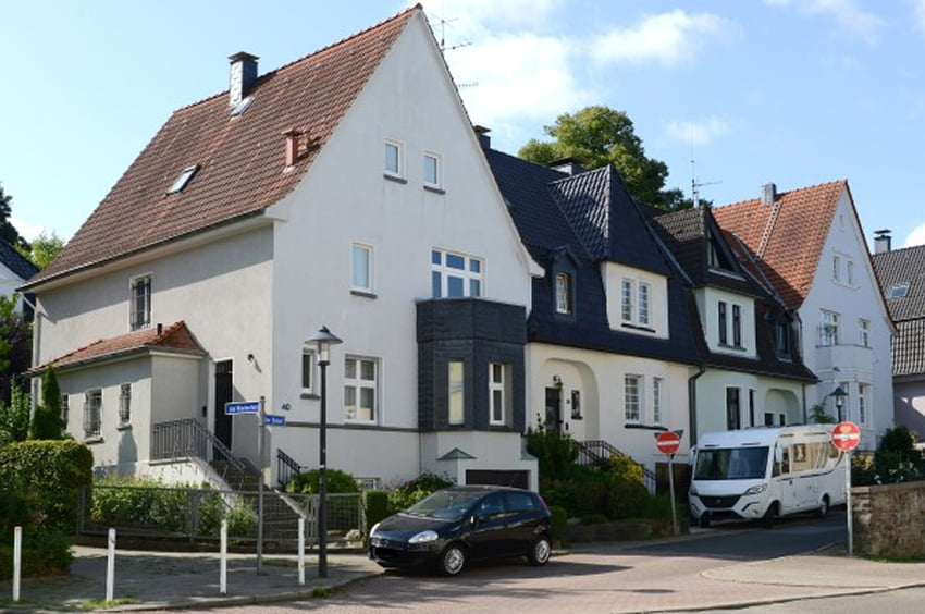 Wohnhäuser mit hellen Fassaden und roten Ziegeldächern an einer ruhigen Wohnstraße.