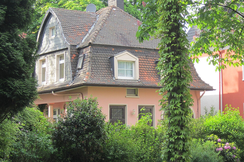 Wohnhaus mit hellem Fassadenanstrich, Ziegeldach und dichtem Gartenbewuchs im Vordergrund.