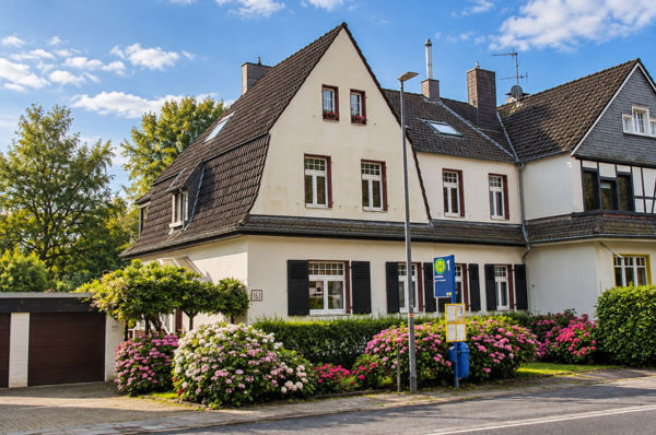 Helles Wohnhaus mit Mansarddach, dunklen Fensterläden und blühenden Hortensien im Vorgarten an einer Bushaltestelle in Essen-Werden.